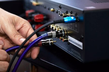 Blå og røde RCA-kabler tilsluttet bagpå en sort elektronisk enhed, hånd holder kablerne
