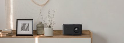 Proiettore con custodia in plastica opaca nera, obiettivo a destra, vista frontale, su credenza in legno chiaro con cornice e vasi