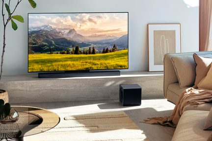 Televisore a schermo piatto nero con cornice ultrasottile in metallo e vetro, visto frontalmente, posizionato su un mobile con soundbar rettangolare nera opaca abbinata e subwoofer cubico liscio posizionato a terra