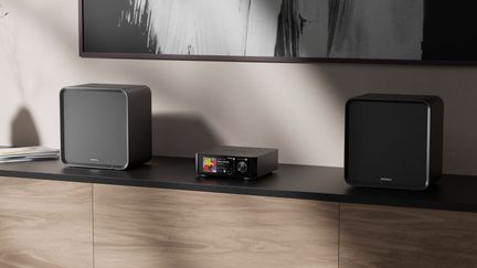 Compact hifi-keten met zwarte versterker met kleurendisplay en twee zwarte vierkante luidsprekers op een houten tv-meubel
