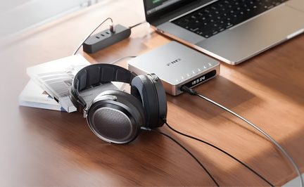 Black and gray circumaural headphones made of brushed metal and plastic, with open-back ear cups and a padded headband, shown from an oblique top view on a wooden desk and connected by cable to a silver audio unit