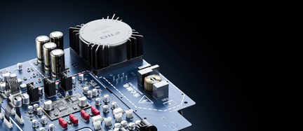 Háromnegyed szögből látható kék PCB anyagú elektronikus kártya, nagy fekete, bordázott hengeres hűtőtesttel, több metalizált elektrolit kondenzátorral, fehér csatlakozókkal és egyéb felületre szerelt apró komponensekkel