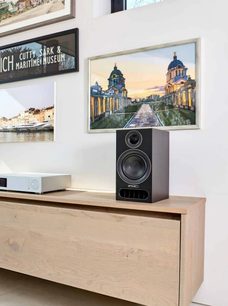 Ηχείο βιβλιοθήκης PMC Prodigy1 μαύρο σε έπιπλο από ανοιχτόχρωμο ξύλο, με δύο κορνιζαρισμένους πίνακες στον τοίχο που απεικονίζουν κτίρια