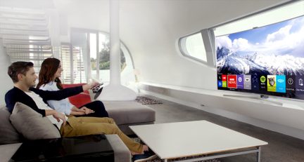 Pareja mirando una pantalla de televisión que muestra un paisaje nevado, sentados en un sofá gris en una sala de estar moderna