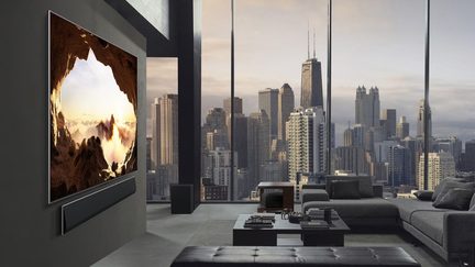 Stor sort vægmonteret tv med slank metalramme og ultratyndt fladskærmsdisplay, ledsaget af en matchende sort rektangulær soundbar, set fra en trekvart sidevinkel