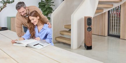 Svetlá drevená hi-fi stĺpová reprosústava v modernom obývacom pokoji, pri páre, ktorý si prezerá knihu pri veľkom stole