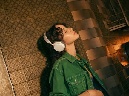 En ung kvinde med brunt krøllet hår, iført en grøn denimjakke, lytter til musik med hvide hovedtelefoner