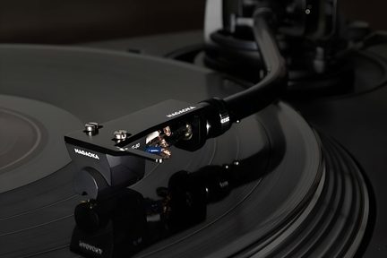 Skråt nærbillede af en mat sort metal pickup og headshell med Nagaoka-inskription, sølvfarvede skruer og synlige farvede ledninger for enden af en buet arm placeret på en sort vinylplade