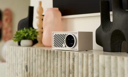 Stříbrný stolní audio přístroj z kovu, přední panel s diagonální mřížkou, kruhový displej a tlačítko, pohled ze tří čtvrtin