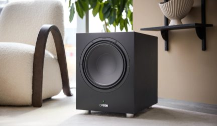 Sort, kubeformet Canton-subwoofer med frontvendt, cirkulær højttaler, placeret i moderne stue på lyst gulv