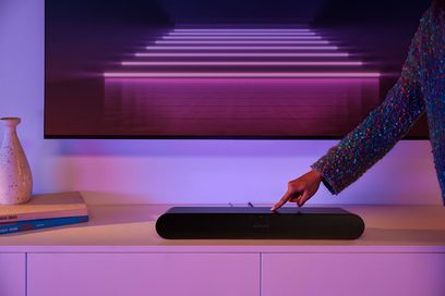 Sonos Ray svart lydplanke, hånd som berører enheten, lilla lys, hvit vegg, bøker, vase