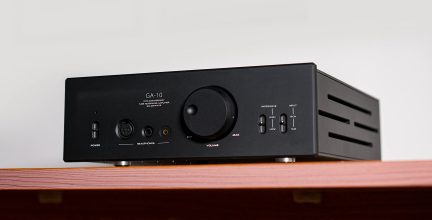 Amplificatore nero, vista frontale, manopola centrale, prese per cuffie, interruttori, testo "GA-10", posizionato su superficie in legno