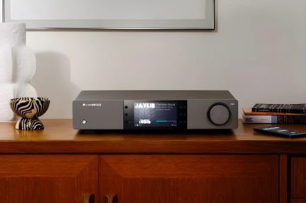 Šedý audio zesilovač s centrálním displejem, kruhovým tlačítkem vpravo, položený na dřevěném nábytku, bílá váza, pruhovaná mísa, knihy