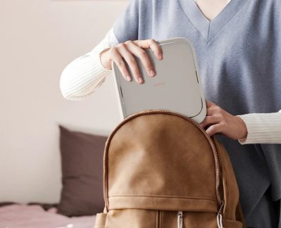 Iemand stopt een lichtgrijs rechthoekig apparaat in een bruine rugzak, vooraanzicht, handen zichtbaar, blauwe kleding