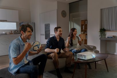 Tre personer sidder på en grå sofa i en stue og ser entusiastisk og glad på en projektionsskærm
