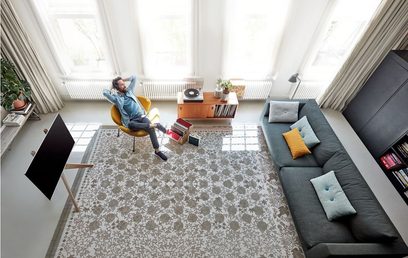 En mand sidder i en gul lænestol i en moderne stue med en grå sofa, et tæppe med mønstre og store vinduer