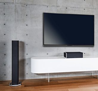 Definitive Technology BP9040 sort søjlehøjttaler, soundbar og basreflekshøjttaler, på hvidt TV-møbel, betonvæg