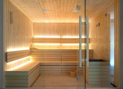 Indvendigt billede af en sauna i lyst træ med horisontale bænke, vægge med træpaneler, integreret belysning, glasdør og en træspand