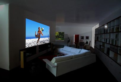 Mørkt interiør med en skærm, der projicerer et billede af en kvinde i badedragt på en solrig strand, med en hvid sofa foran