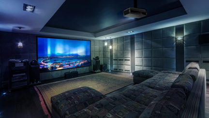 Sala cinema con schermo a parete luminoso, divano in tessuto a motivi, tappeto centrale, pareti imbottite e proiettore a soffitto