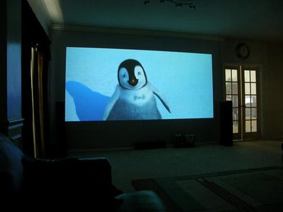 Projektion af en tegnefilmspingvin på en stor skærm i et mørkt rum med vinduer og døre