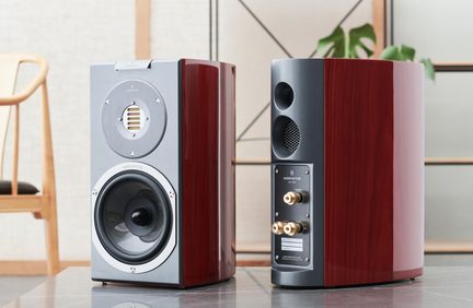 Et par Audiovector reolhøjttalere i rød lakeret træ, antracitgrå metalfronter med synlig rektangulær bånddiskant og cirkulær basenhed på den ene, den anden viser bagsiden med to runde basreflekshuller, et gitter og forgyldte terminaler, set forfra og bagfra på et bord