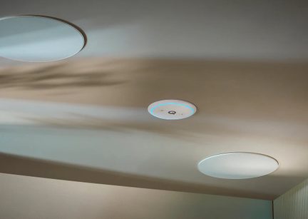 View from below of a beige ceiling with three recessed white discs: two with perforated metal circular grilles, and a smaller central module in smooth plastic featuring a blue light ring and pictograms