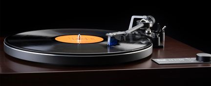 Toca-discos de vinil visto em três quartos de frente, base em madeira marrom envernizada, prato preto com um disco de etiqueta laranja, braço metálico cinza com cápsula azul na extremidade e botão de ajuste preto no canto direito