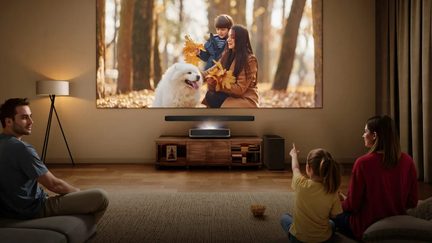 Moderní béžový salón, černý matný ultra krátkohledý videoprojektor umístěný na nízkém nábytku ze světlého dřeva pod širokým promítaným obrazem, pohled zepředu