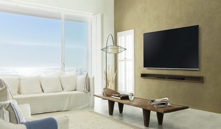 Svetainė su balta sofa, medine žurnaliniu staliuku, stovo lempa, sienine televizija, langu ir baltais užuolaidėlėmis