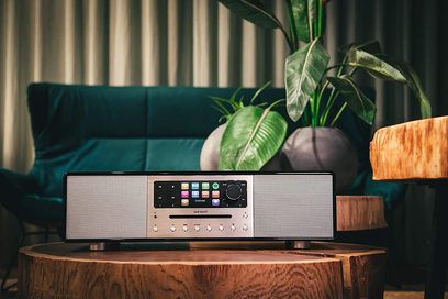 Sonoro Meisterstück digital radio i blank sort og sølv, sort front med skærm og knapper, placeret på en træstamme med grøn plante bagved