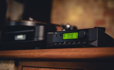 Juodas metalinis garso įrenginys, matomas iš trijų ketvirčių priekio, žalias LCD ekranas, garso reguliavimo mygtukas, eilė mygtukų, ant medinio baldo