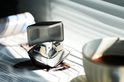 Sølvfarvede trådløse høretelefoner i et åbent etui på et bord, naturligt lys, hvid kop til højre, persienner i baggrunden