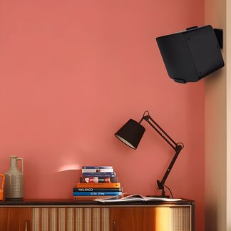Altoparlante da parete nero, lampada da scrivania articolata nera, pila di libri colorati, caraffa grigia, muro rosa, mobile in legno