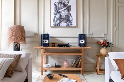 Vooraanzicht van een woonkamer met houten tafel, platenspeler, 2 luidsprekers, lamp met rieten kap, bloemenvaas, schilderij