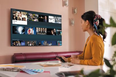 Samsung QE75Q60T 2020 televisie met gebruikersinterface met miniaturen van films en series, zijwaarts gezien, met een vrouw die ervoor zit