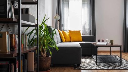 Modern salon, gri köşe koltuk, sarı yastıklar, yeşil bitki, kitaplık, siyah sehpa, geometrik halı ve büyük pencereler