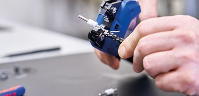 Detailní záběr na ruce osoby držící a používající modrý elektrický nástroj k řezání nebo vrtání do neviditelného předmětu.