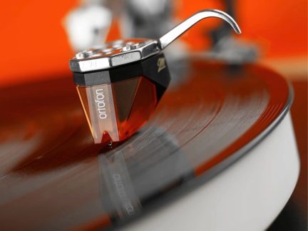 Cellule phono en plastique orange translucide et noir, corps en métal argenté, vue macro latérale sur disque vinyle rainuré