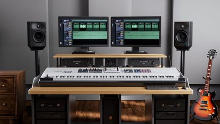 Frontalt vue af et lydstudie: skrivebord i lyst træ, to sorte skærme og højttalere, stort hvidt keyboard, sunburst-guitar
