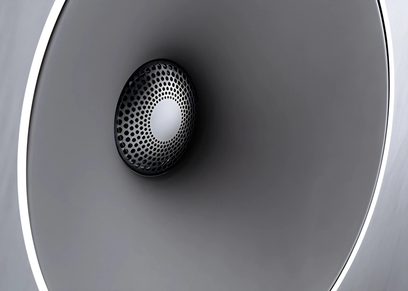 Black circular speaker seen from the front, integrated into a curved dark gray surface, illuminated by a white light