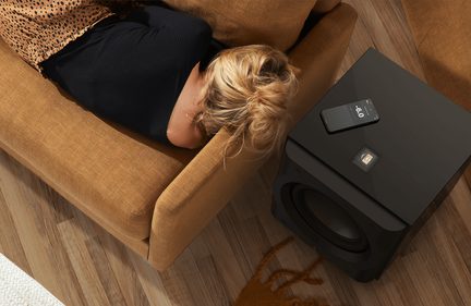 Vedere de sus, canapea maro, persoană întinsă, păr blond, bluză neagră, subwoofer negru, smartphone cu ecranul aprins