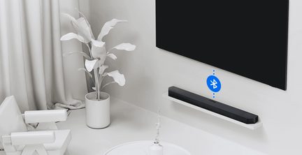 Svart soundbar i texturerad plast, sedd i trekvarts vinkel placerad på en vit vägghylla under en svart platt-TV, med blå Bluetooth-ikon som visar anslutning