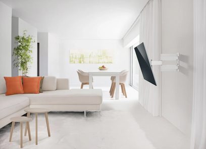 Suporte de TV de parede branco fixado na parede de uma sala de estar moderna com sofá bege, almofadas laranja, mesa de centro de madeira clara e planta verde