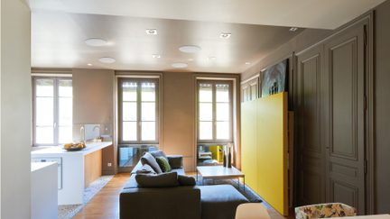 Vista frontale di un soggiorno con parquet in legno chiaro e cucina a vista, dotato di un divano angolare grigio in tessuto, un'isola bianca laccata con rubinetto, un pannello di stoccaggio giallo opaco e due ampie finestre incorniciate da boiserie color taupe.