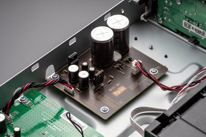 Vihreä piirikortti, jossa on elektroniikkakomponentteja, kaksi isoa hopeista kondensaattoria ja punaisia ja mustia kaapeleita, lähikuva