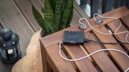 ЦАП-підсилювач для навушників Chord Mojo 2 чорного кольору з сірою парою внутрішньоканальних навушників на дерев’яній лавці надворі