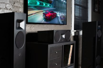 A screen displaying a red car in motion, two large black speakers, a black cabinet with drawers and vinyl records, a brick wall