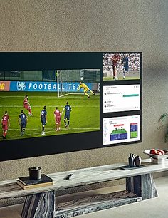Skærm der viser en fodboldkamp med spillere på en grøn bane, som ser ud til at være i et kontrolrum eller et visningsrum