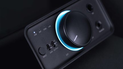 Close-up three-quarter view of the front face of a rectangular black matte metal audio unit, featuring a large knob surrounded by a blue LED light, input indicators, two switches, and jack sockets